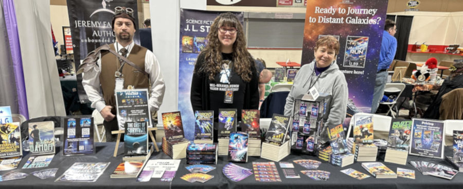 Authors Jeremy Fabiano, J. L. Stowers, and LJ Dix standing behind their table of fantasy and science fiction books at 2024 Gem State Comic Con in Boise, Idaho.